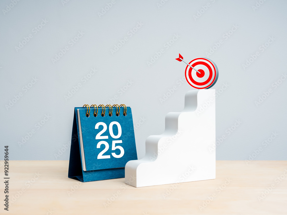 Wall mural Happy new year 2025 background. 2025 numbers on blue small desk calendar near white stairway with 3d target icon on top on wood table, white background. Business growth, goal and success plan concept.