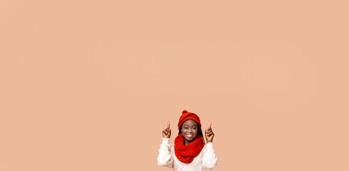 A cheerful young African American woman with a big smile is wearing a cozy red hat and matching scarf. She is enthusiastically pointing upward at copy space