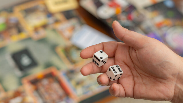 Woman's hand holding playing dices. Throw Two Dice On a Table With Board game. Hand throws the dice on the background of colorful blurred fantasy Board games, gaming moments in dynamics