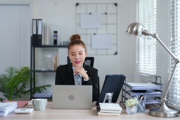 A professional woman in a black suit is sitting at her desk, concentrating on her work with a...