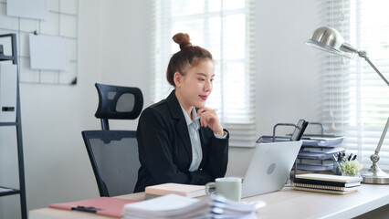 A professional woman in a black suit is sitting at her desk, concentrating on her work with a laptop, surrounded by files and office supplies, showing a focused work atmosphere