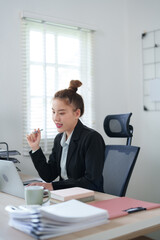 A professional woman in a black suit is sitting at her desk, concentrating on her work with a laptop, surrounded by files and office supplies, showing a focused work atmosphere