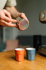 Barista sprinkling cocoa powder into two colorful cups of coffee on a wooden table.
