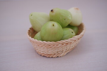 A small wicker basket filled with light green water guava sits on a pale wooden surface. The fruit has a glossy, slightly bumpy texture and a conical shape.