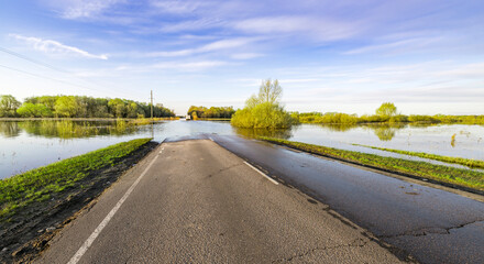 A tranquil rural road is partially submerged as floodwaters rise, contrasting with the greenery and blue sky above, depicting nature's power in the quiet of springtime.