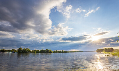 A serene river flows under a sky filled with fluffy clouds. The golden light filters through, casting a glow on the calm water. Trees on the opposite bank complete the beautiful scene