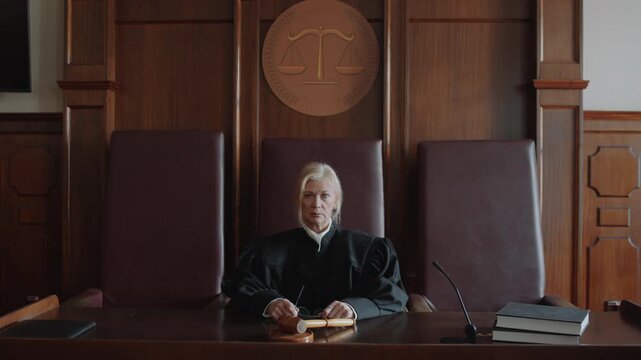 Portrait of Caucasian mature serious female judge dressed in black mantle sitting at bench and posing for camera in courtroom