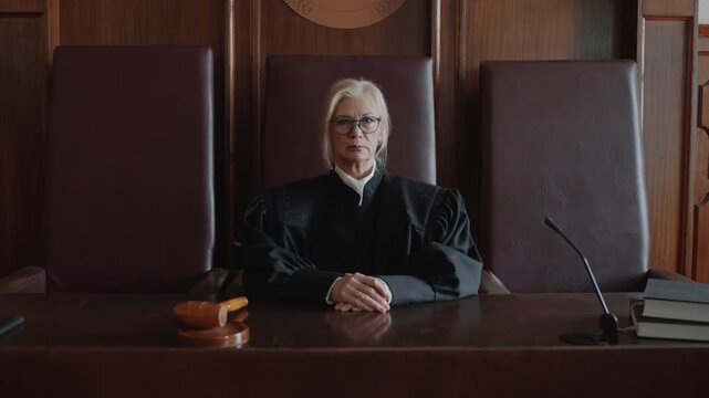 Zoom in portrait of Caucasian mature impartial female judge wearing glasses and black mantle looking at camera while sitting at wooden desk in courtroom