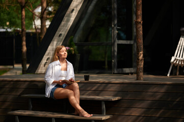 Young blonde woman sits on the veranda of her house making notes in a notebook on a warm summer evening. Middle-aged European woman sitting on the stairs of her house making notes in her diary.