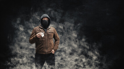 Drug dealer, dangerous hooded criminal man in black mask and brown leather jacket holding transparent plastic bag with drugs, on a dark background with smoke