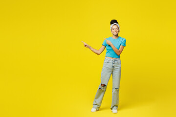Fototapeta premium Full body young smiling happy fun Latin woman she wears blue top t-shirt headband casual clothes point index finger aside on area mock up isolated on plain yellow background studio. Lifestyle concept.