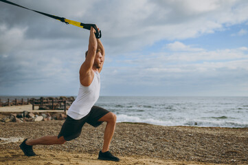 Sideways young strong sporty toned fit sportsman man in sports clothes use trx loops do exercise do...