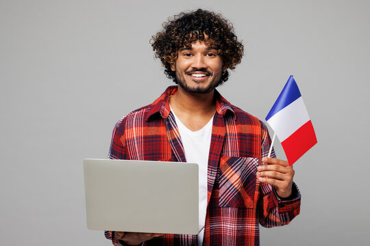 Young smart happy smiling Indian IT man he wearing red shirt casual clothes hold French flag use work on laptop pc computer isolated on plain grey color background studio portrait. Lifestyle concept.