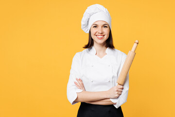 Young smiling happy chef cook baker woman wear white shirt black apron uniform toque chefs hat hold in hand rolling pin looking camera isolated on plain yellow background studio. Cooking food concept.