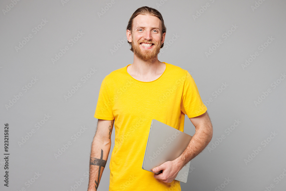 Wall mural Smiling happy fun cheerful young IT man wear yellow t-shirt casual clothes hold in hand closed laptop pc computer looking camera isolated on plain grey background studio portrait. Lifestyle concept.