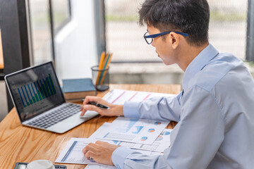 Young businessman working on his project in his office checking business result data from the printed sheets and his laptop computer