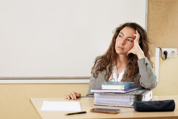 Woman overwhelmed by work overload at the table in her office or high school classroom, she looks to the side thinking where to start.