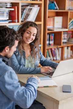 The woman searches for information on the laptop and the man stops listening to music on his headphones to help her study with her notebook or to do her college work.