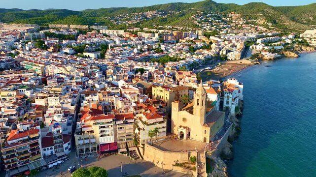 Orbiting shot of Sitges coastal village and Iglesia de San Bartolome at sunset, holiday in Spain, drone view of Spanish mediterranean village