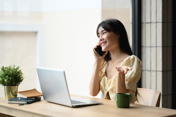 Charming young asian woman talking on smart phone while looking away at her workplace