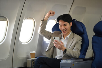 Happy businessman sitting in an airplane seat, looking at his smartphone and celebrating with a joyful expression