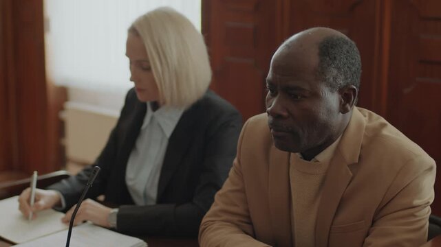 Nervous African American accuser thinking about case, then sharing his thought with Caucasian female prosecutor sitting next to him in courtroom