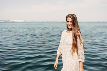 A woman is walking on the beach wearing a dress. The water is calm and the sky is clear.