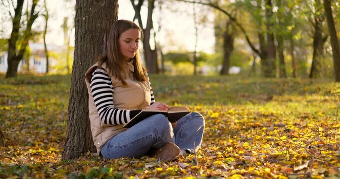 Woman reading a book while sitting under a tree in an autumn park