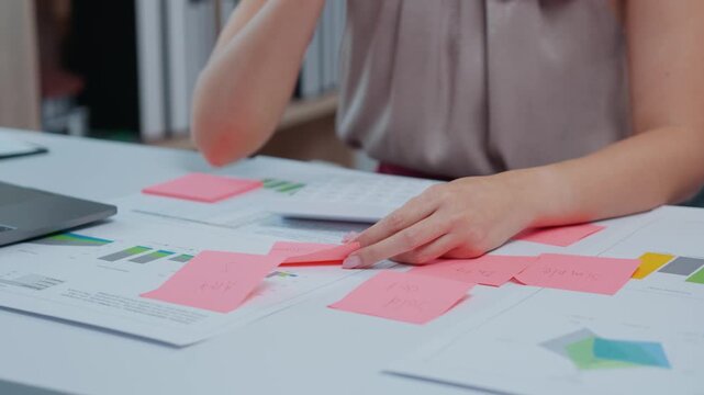 Person working at a desk with charts, graphs, and sticky notes, organizing and analyzing data, with a laptop in view. The focus is on hands moving papers and writing