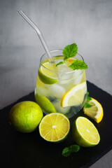 Glass with soda, ice, mint, lemon and lime slices next to cut citrus fruits and ice cubes on a black tray on a gray background