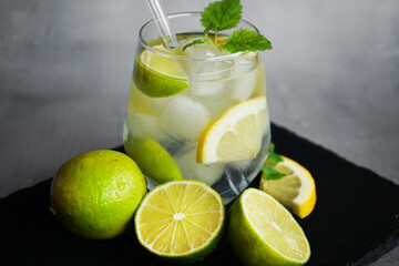 Glass with soda, ice, mint, lemon and lime slices next to cut citrus fruits and ice cubes on a black tray on a gray background