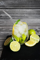 Glass with soda, ice, lemon and lime slices next to cut citrus fruits on a black tray on a gray wooden background