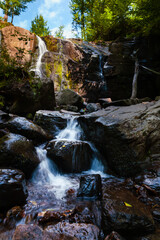 A captivating image of a serene waterfall cascading down a rocky cliff surrounded by lush greenery. The water flows gently over the dark rocks, creating a peaceful and tranquil scene in a forested are
