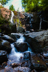 A captivating image of a serene waterfall cascading down a rocky cliff surrounded by lush greenery. The water flows gently over the dark rocks, creating a peaceful and tranquil scene in a forested are