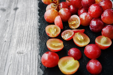 A bunch of large pink grapes next to cut grapes on a black tray on a gray wooden background