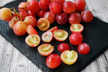 A bunch of large pink grapes next to cut grapes on a black tray on a gray wooden background