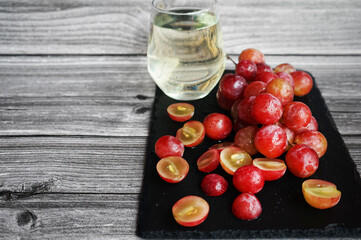 A bunch of large pink grapes next to a glass of white wine on a black tray on a gray wooden background