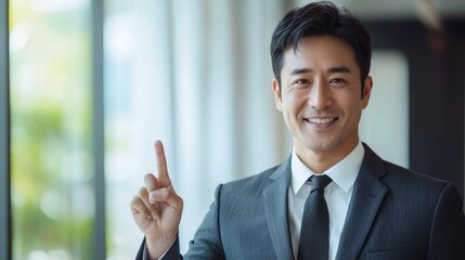 Asian businessman wearing a sleek suit stands confidently in a bright studio, raising his index finger as he smiles. The background is clean and minimal, offering ample copy space for potential use
