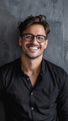 Obraz premium A smiling young man with black hair, glasses and braces poses against a grey background in a casual and relaxed setting. He radiates charm in a black shirt with a deep V-neckline