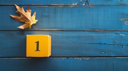 A single autumn leaf and a yellow block with the number 1 sit on a rustic, light-blue wooden surface, evoking a sense of simplicity, playfulness, and the changing seasons.