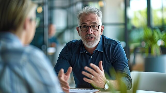 Middle-aged caucasian man with beard gesturing while talking Indoor professional setting Engaged in conversation Warm lighting Networking event