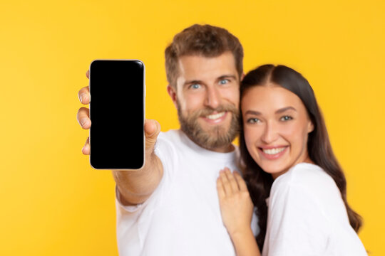 Happy loving European couple embracing and showing display of mobile phone with blank screen, selective focus on cellphone, mockup