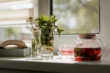 red strawberry and mint tea in a glass teapot, summer mood, still life on the windowsill, chamomile bouquet