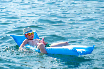 chic child basking in hat and sunglasses enjoys luxurious beach day, young fashionist floating on inflatable raft while sipping refreshing beverage, Childhood opulence in picture-perfect moment