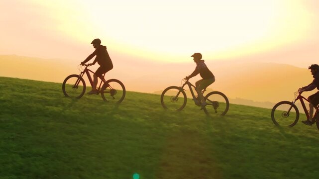 AERIAL: Golden evening sunbeams shine on mountain bikers pedalling up a grassy hill in the beautiful Slovenian mountains. Scenic drone view of active people riding e-bikes in the scenic Dolomites.