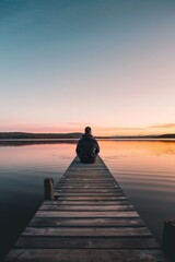 A person sits at the end of a wooden dock at a tranquil lake, watching the beautiful sunset, reflecting on life and enjoying a peaceful moment in nature during the quiet evening time.
