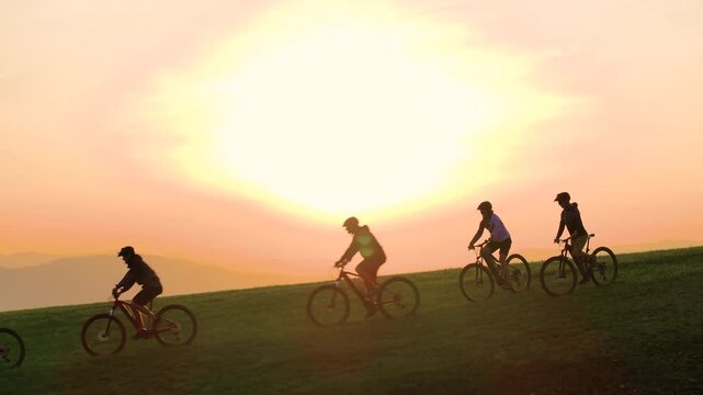 AERIAL, LENS FLARE: Mountain bikers high five before descending into the verdant valley at breathtaking early autumn sunset. Scenic aerial shot of group of friends riding e-bikes in the idyllic hills.
