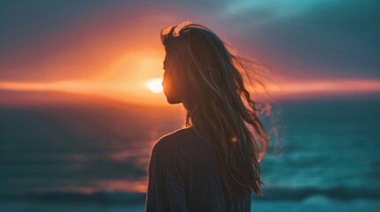 Fototapeta premium A woman stands by the ocean, watching the sunset with her hair flowing in the breeze. The colors of the sunset create a vibrant backdrop, symbolizing tranquility and reflection.