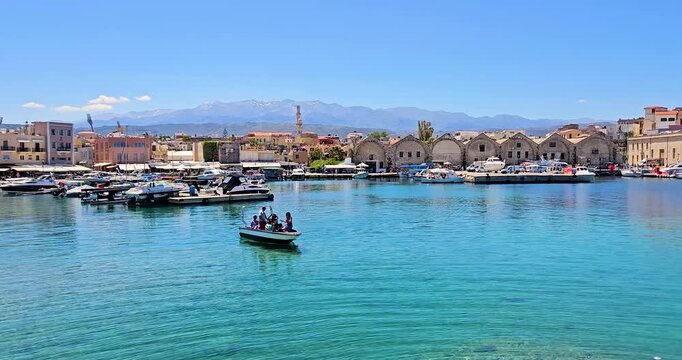 Landscape of Chania Old Port with Venetian shipyards, tourist boats passing and White Mountains in the background. 