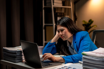 A woman is sitting at a desk with a laptop and piles of papers